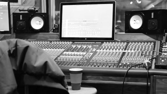 A Black & white photo of a recording studio mixing board, with a coffee cup on a stool in the foreground